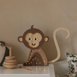 May include: A wooden monkey night light with a brown and beige color scheme. The monkey has a smiling face and a long tail. The light has small, warm white LED lights that illuminate the monkey's body.