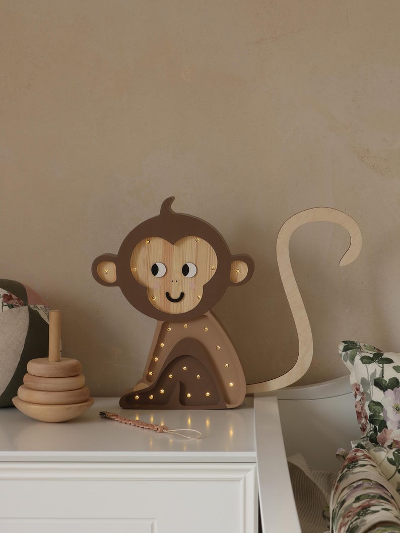 May include: A wooden monkey night light with a brown and beige color scheme. The monkey has a smiling face and a long tail. The light has small, warm white LED lights that illuminate the monkey's body.