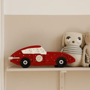 May include: A red wooden toy race car with white accents and black wheels sits on a shelf next to two stuffed animal toys. The car has the number 27 on the side. The stuffed animals are a dog and a bunny.