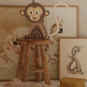May include: A wooden monkey night light with a brown body and a light brown tail. The monkey has a smiling face with black eyes and a brown nose. The night light is sitting on a wooden stool.