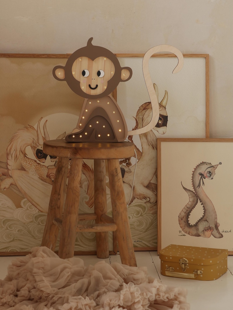 May include: A wooden monkey night light with a brown body and a light brown tail. The monkey has a smiling face with black eyes and a brown nose. The night light is sitting on a wooden stool.