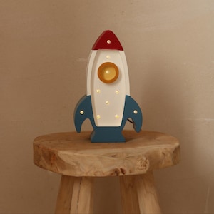 Könnte beinhalten: Eine holzfarbene Raketenlampe mit roter Spitze, weißem Korpus und blauen Flossen. Die Lampe hat ein gelbes kreisförmiges Licht in der Mitte und mehrere kleinere Lichter. Sie steht auf einem kleinen, runden Holzschemel.