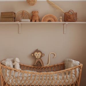 May include: A wicker crib with a white mattress and a stuffed animal. The crib is decorated with a wooden monkey toy and a woven rainbow.