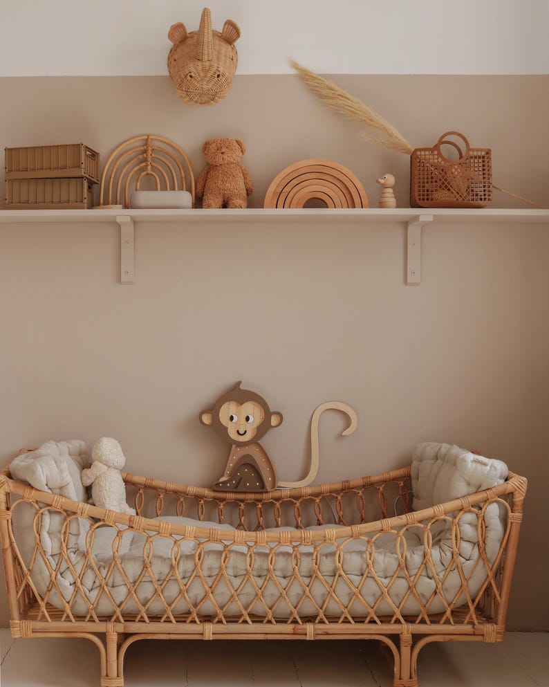 May include: A wicker crib with a white mattress and a stuffed animal. The crib is decorated with a wooden monkey toy and a woven rainbow.