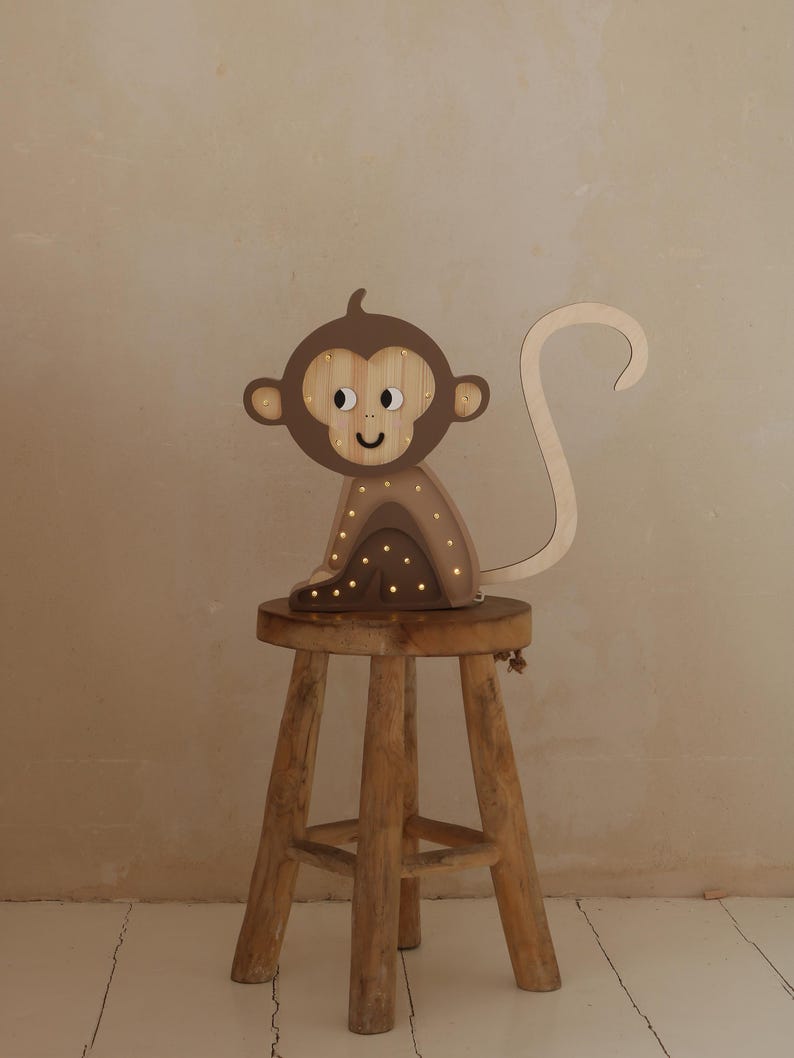 May include: A wooden monkey night light with a brown finish and a smiling face. The light is sitting on a wooden stool.
