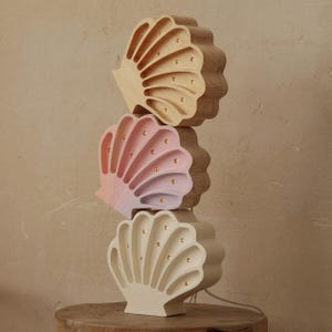 May include: Three seashell-shaped lamps stacked on a wooden surface. The lamps are in natural wood, pink and beige, each with small golden lights. The lamps are connected by a white cord.