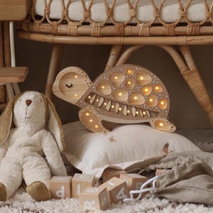 Puede incluir: Una luz de noche de madera con forma de tortuga con luces LED cálidas, apoyada sobre una almohada blanca. Un conejito de peluche y bloques de madera con letras están cerca. La escena sugiere un cuarto de niños.