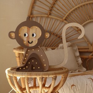 May include: A wooden monkey night light with a brown body and a light brown face. The monkey has a long tail and is sitting on a small, round table. The night light has small, white lights that illuminate the monkey's face.
