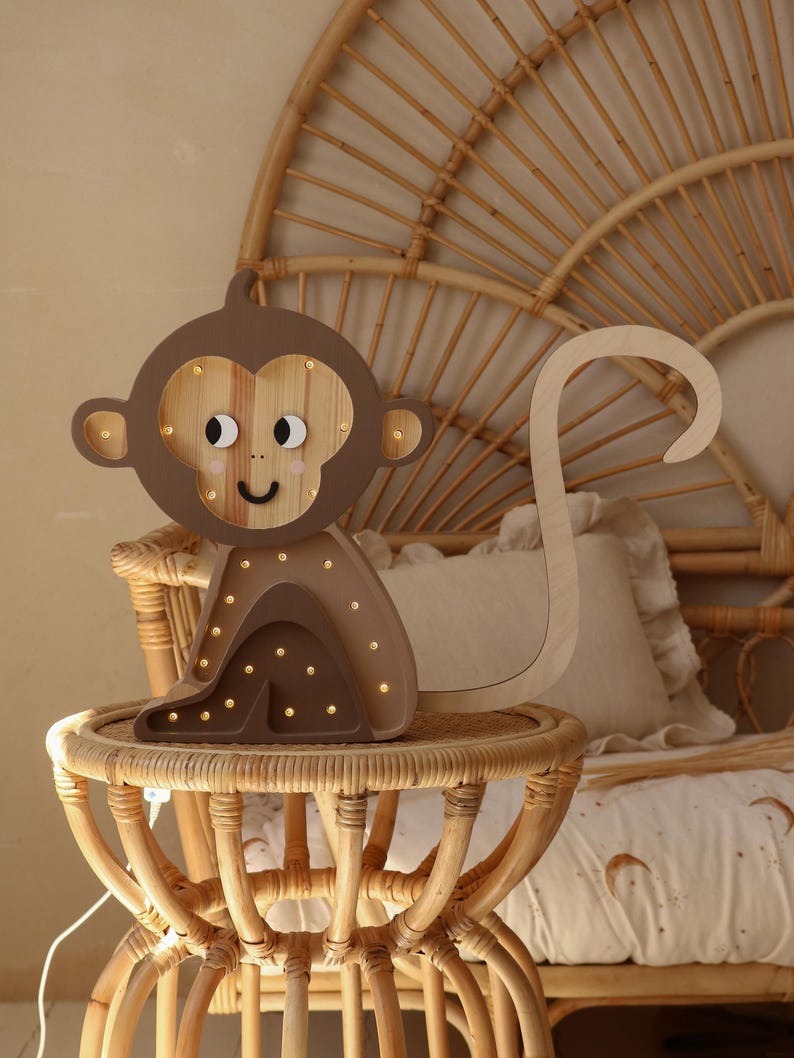 May include: A wooden monkey night light with a brown body and a light brown face. The monkey has a long tail and is sitting on a small, round table. The night light has small, white lights that illuminate the monkey's face.