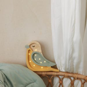Könnte beinhalten: Eine hölzerne Vogel-Nachtlampe mit gelbem Körper und grünen Flügeln. Der Vogel hat ein kleines, rundes Gesicht mit einem Lächeln und ein paar kleinen, weißen Punkten für Augen. Das Licht ist an und die Punkte leuchten.