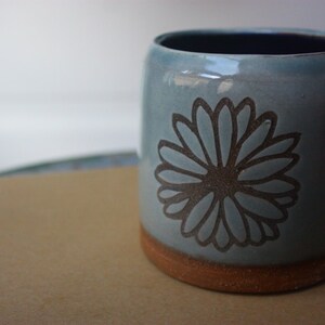 May include: A light blue ceramic mug with a brown floral design. The mug has a brown rim and base.