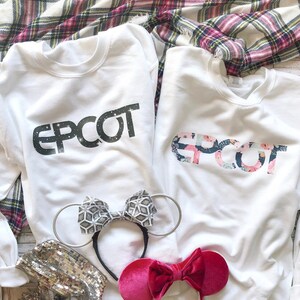 May include: Two white sweatshirts with the word "EPCOT" printed on them. One sweatshirt has the word in black with a glittery effect, and the other has the word in a floral pattern. There are two pairs of Minnie Mouse ears, one with a silver bow and the other with a pink bow.
