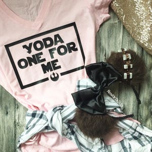 K&ouml;nnte beinhalten: Rosa V-Ausschnitt-T-Shirt mit schwarzem Text, der "Yoda One For Me" innerhalb eines schwarzen Quadrats lautet. Das Shirt liegt auf einer Holzoberfl&auml;che mit einem schwarz-wei&szlig; karierten Hemd und einem braunen, flauschigen Stirnband mit einer schwarzen Schleife.