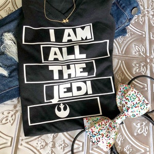 May include: Black t-shirt with white text that reads "I AM ALL THE JEDI" with a Rebel Alliance symbol below the text. The shirt is laying on a white surface with a pair of black mouse ears with a white bow with colorful confetti.