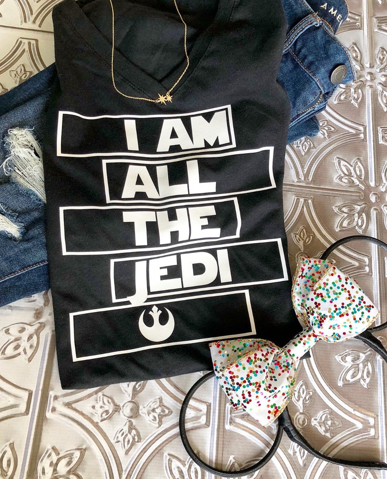 May include: Black t-shirt with white text that reads "I AM ALL THE JEDI" with a Rebel Alliance symbol below the text. The shirt is laying on a white surface with a pair of black mouse ears with a white bow with colorful confetti.