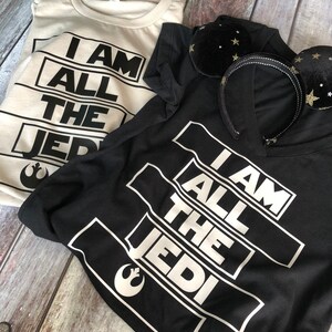 May include: Two t-shirts with the text "I am all the Jedi" printed on them. One shirt is black and the other is white. Both shirts feature the Star Wars Rebel Alliance symbol.