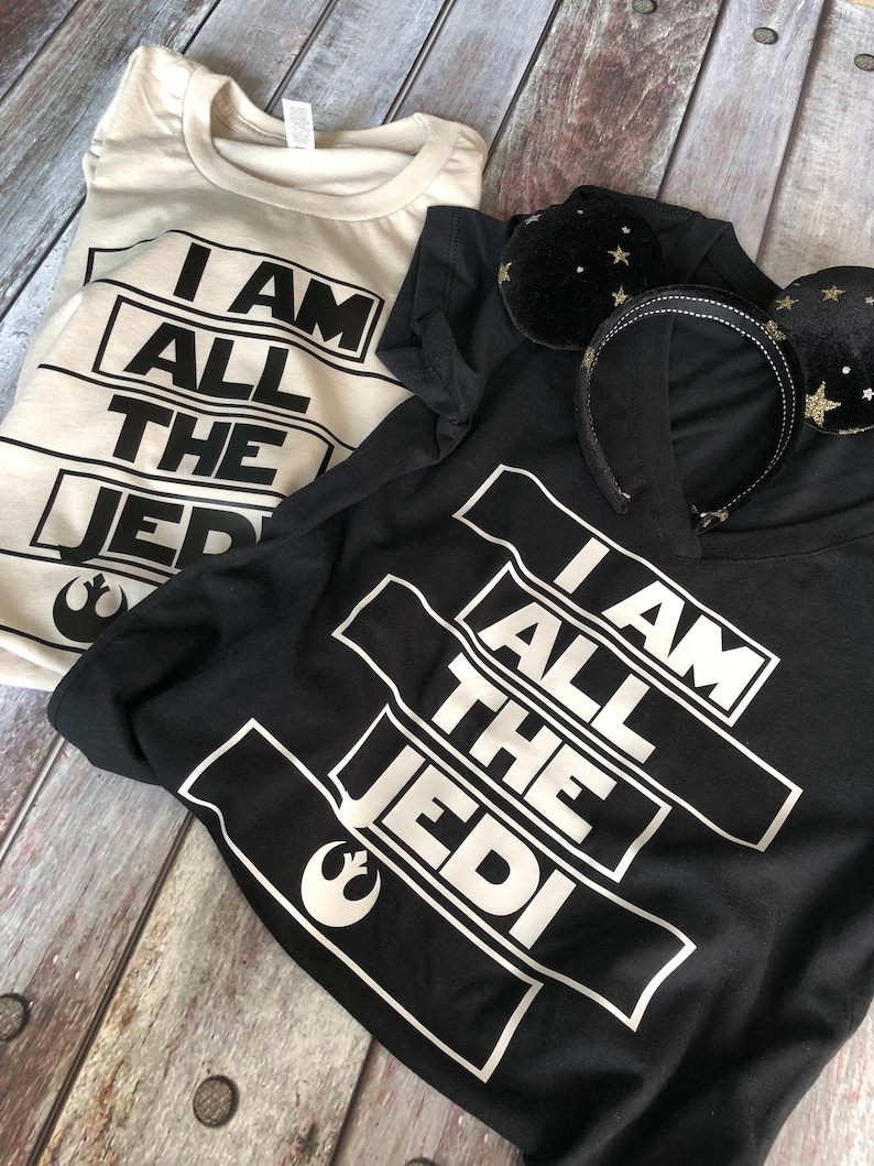 May include: Two t-shirts with the text "I am all the Jedi" printed on them. One shirt is black and the other is white. Both shirts feature the Star Wars Rebel Alliance symbol.