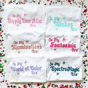 以下が含まれることがあります： カラフルなテキストデザインが施された白いTシャツが5枚。シャツのテキストは「In My Happily Ever After Era」、「In My Wishes Era」、「In My Illuminations Era」、「In My Fantasmic Era」、「In My World of Color Era」です。