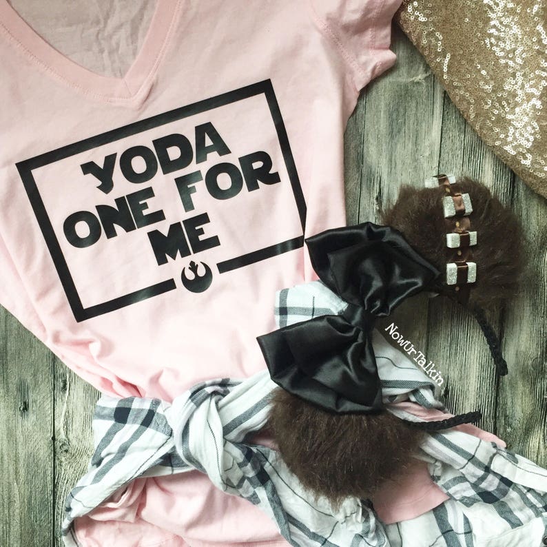K&ouml;nnte beinhalten: Rosa V-Ausschnitt-T-Shirt mit schwarzem Text, der "Yoda One For Me" innerhalb eines schwarzen Quadrats lautet. Das Shirt liegt auf einer Holzoberfl&auml;che mit einem schwarz-wei&szlig; karierten Hemd und einem braunen, flauschigen Stirnband mit einer schwarzen Schleife.