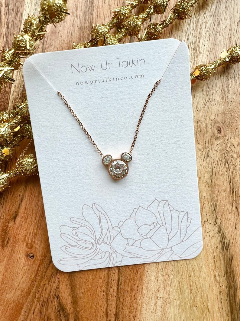 K&ouml;nnte beinhalten: Eine ros&eacute;goldene Halskette mit einem klaren Kristall-Mickey-Mouse-Anh&auml;nger. Die Halskette wird auf einer wei&szlig;en Karte mit dem Text "Now Ur Talkin" und "nowurtalkinco.com" pr&auml;sentiert.