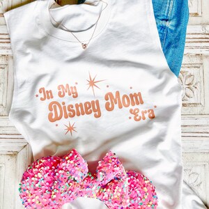 以下が含まれることがあります： 白いノースリーブタンクトップに、星形のローズゴールドの文字で「In My Disney Mom Era」と書いてあります。ピンクのキラキラしたミニーマウスのヘッドバンドがタンクトップの上にあります。