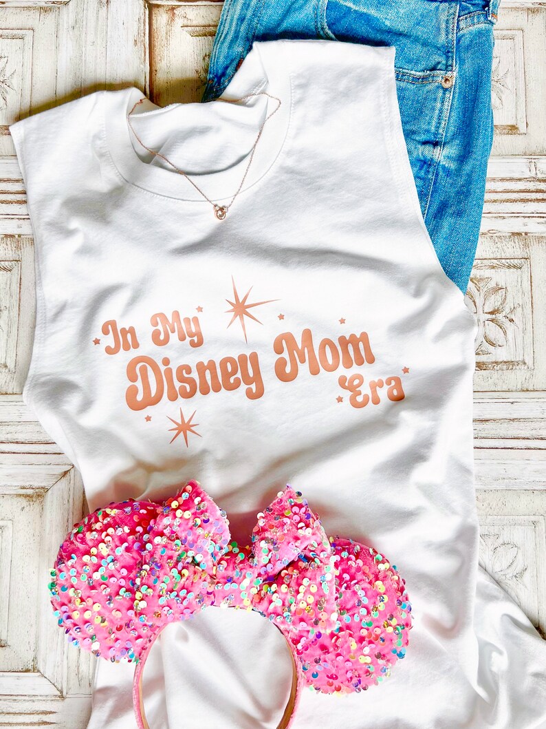 以下が含まれることがあります： 白いノースリーブタンクトップに、星形のローズゴールドの文字で「In My Disney Mom Era」と書いてあります。ピンクのキラキラしたミニーマウスのヘッドバンドがタンクトップの上にあります。