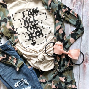 May include: A beige t-shirt with the text "I AM ALL THE JEDI" in black block letters. The shirt is paired with a camouflage cardigan, distressed blue jeans, and a pink bow headband.