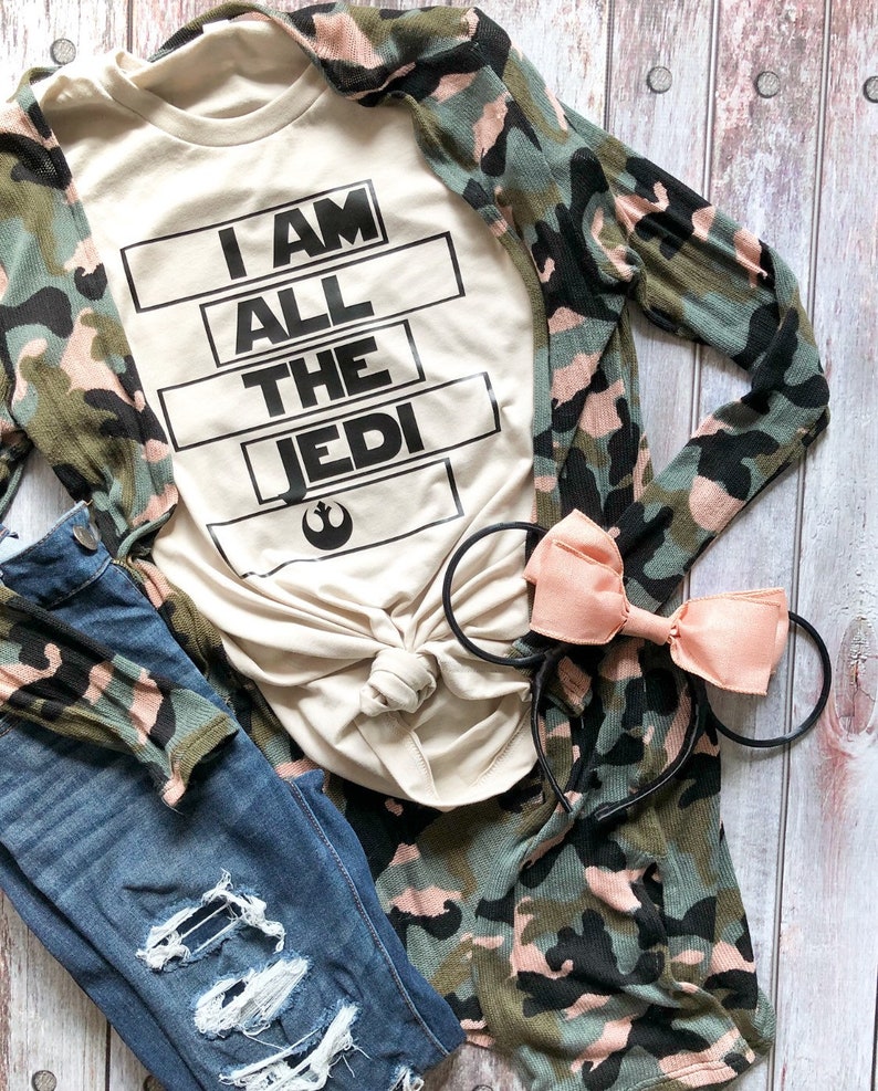 May include: A beige t-shirt with the text "I AM ALL THE JEDI" in black block letters. The shirt is paired with a camouflage cardigan, distressed blue jeans, and a pink bow headband.