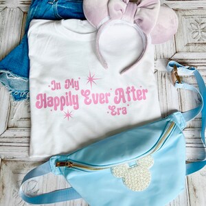 以下が含まれることがあります： ピンクの文字で「In My Happily Ever After Era」と書かれた白いTシャツ。パールで飾られたミッキーマウスの頭のデザインが施された薄いブルーのウエストバッグ。薄いパープルのベルベット製のヘッドバンドにリボンが付いている。
