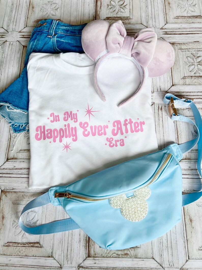 以下が含まれることがあります： ピンクの文字で「In My Happily Ever After Era」と書かれた白いTシャツ。パールで飾られたミッキーマウスの頭のデザインが施された薄いブルーのウエストバッグ。薄いパープルのベルベット製のヘッドバンドにリボンが付いている。