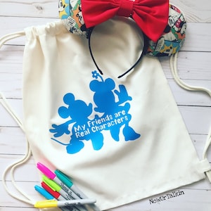 May include: A white drawstring bag with the text "My Friends are Real Characters" and a blue silhouette of Mickey Mouse and Minnie Mouse. The bag is next to a red bow headband with a Mickey Mouse print and a set of colorful markers.