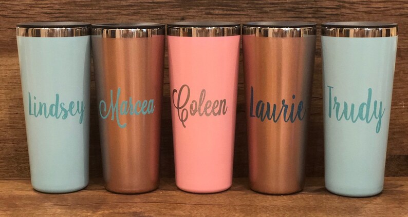 May include: Five stainless steel travel mugs with personalized names: Lindsey, Marcea, Coleen, Laurie, and Trudy. The mugs are in various colors: light blue, copper, pink, and teal.