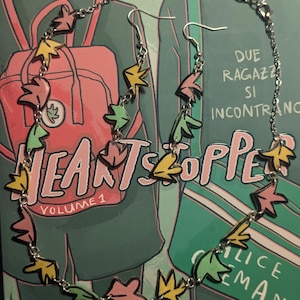 Może przedstawiać: Srebrny naszyjnik i kolczyki z kolorowymi zawieszkami w kształcie liści. Naszyjnik i kolczyki są pokazane na okładce książki z tytułem "Heartstopper" i tekstem "Due Ragazzi Si Incontrano".