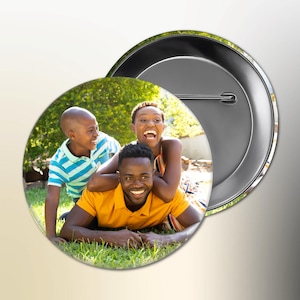 Könnte beinhalten: Ein runder Ansteckbutton mit einer Familie von drei Personen, die auf einer Wiese lachen und lächeln. Der Button hat eine glänzende Metallrückseite und eine Sicherheitsnadel.