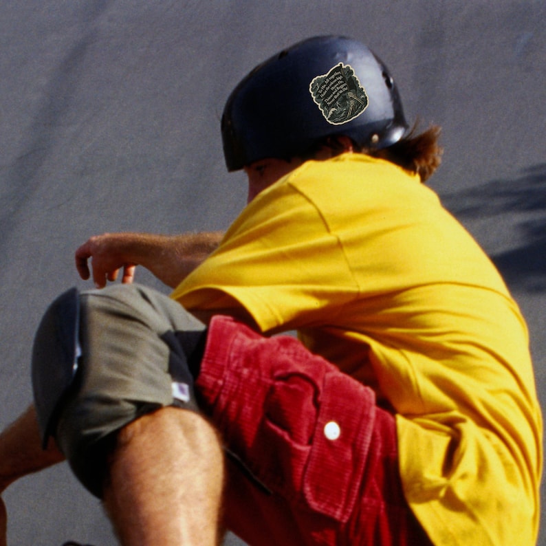 May include: A person wearing a black helmet with a sticker, a yellow t-shirt, red corduroy shorts, and gray knee pads. The person is sitting down, possibly skateboarding or rollerblading.
