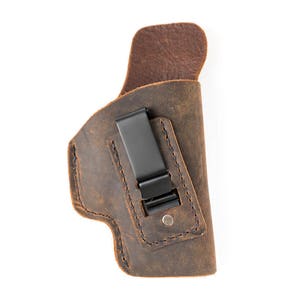 May include: A brown leather holster with a black metal clip. The holster has a stitched design and is designed to hold a firearm.