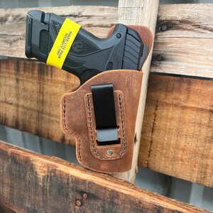 Puede incluir: Una funda de cuero marr&oacute;n con un clip negro para una pistola. La funda est&aacute; unida a una superficie de madera. La pistola es una Ruger Lite Rack Security-380 negra con una pegatina amarilla que dice "Ruger Lite Rack Security-380".