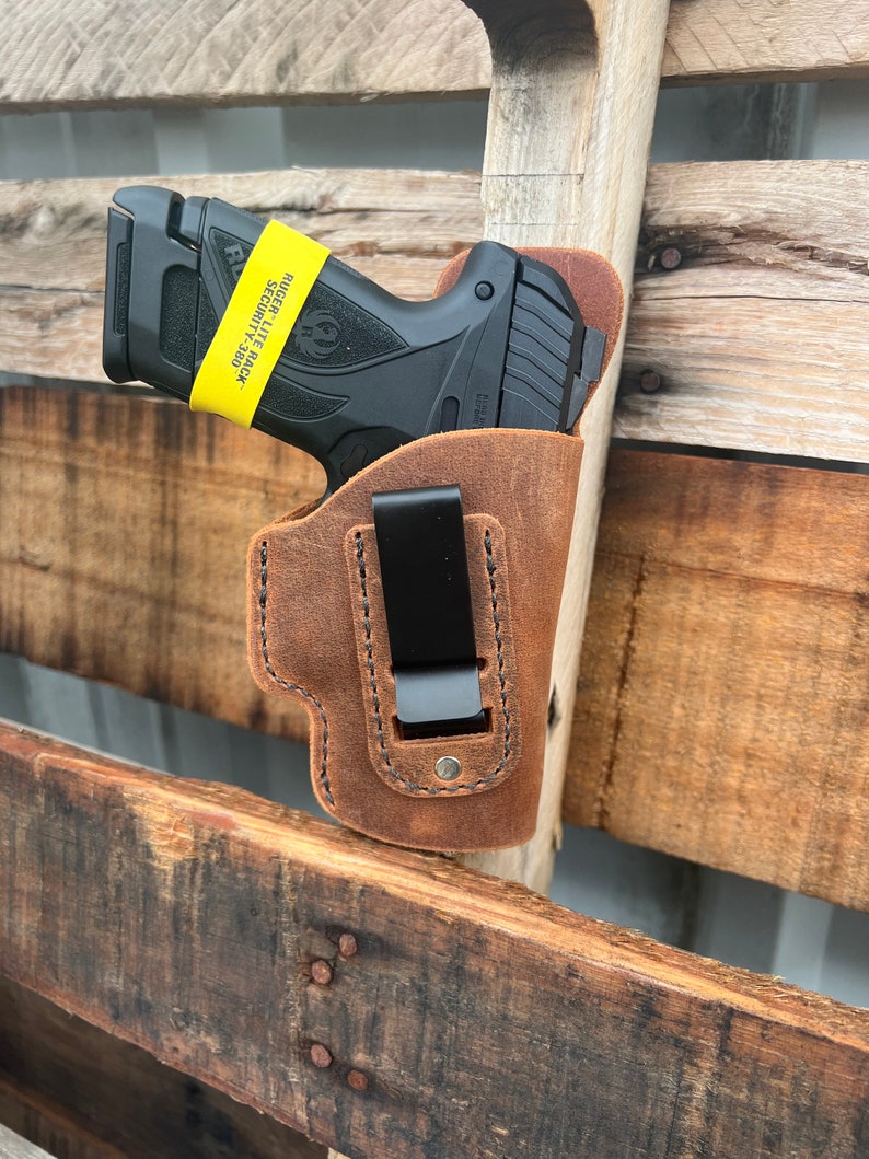 Puede incluir: Una funda de cuero marr&oacute;n con un clip negro para una pistola. La funda est&aacute; unida a una superficie de madera. La pistola es una Ruger Lite Rack Security-380 negra con una pegatina amarilla que dice "Ruger Lite Rack Security-380".