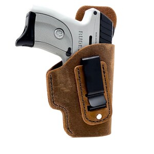 May include: A brown leather holster with a black clip for a silver handgun. The gun has the word "RUGER" printed on the side.