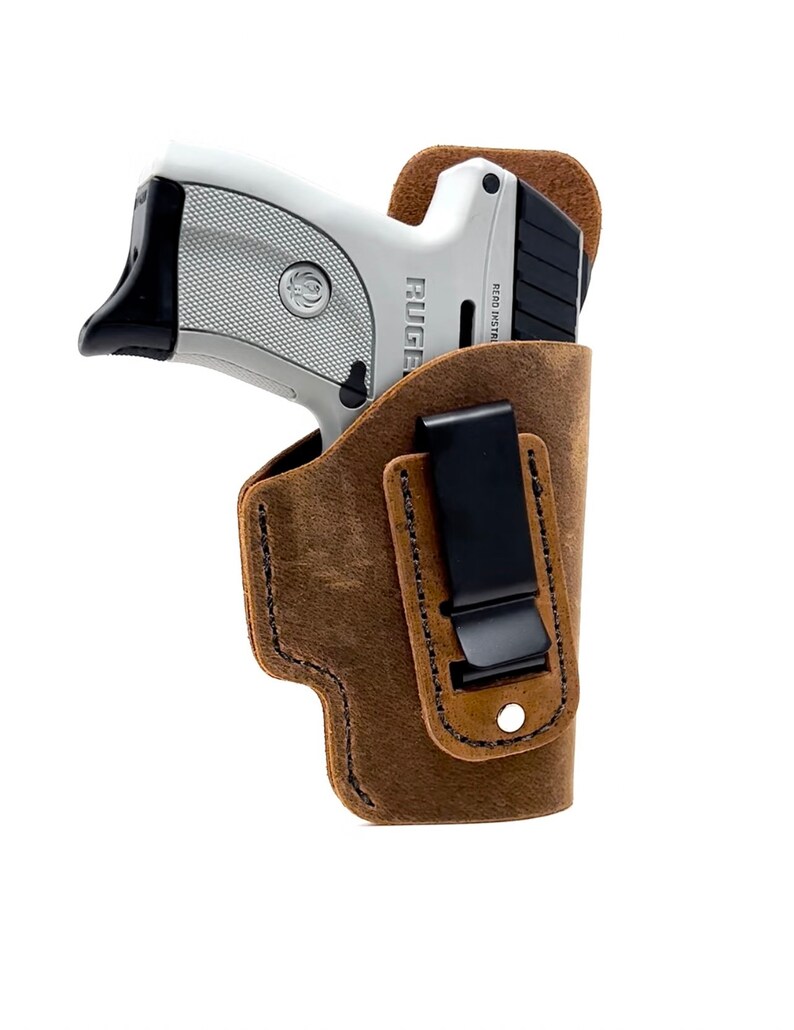 May include: A brown leather holster with a black clip for a silver handgun. The gun has the word "RUGER" printed on the side.