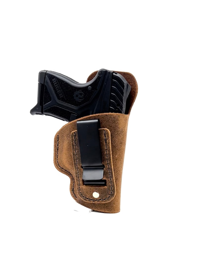 Puede incluir: Una funda de cuero marr&oacute;n con un clip para llevar una peque&ntilde;a pistola negra. La pistola tiene la palabra "RUGER" impresa en el lateral.