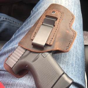 May include: A brown leather holster with a metal clip, holding a black handgun. The holster is worn on a pair of blue jeans.