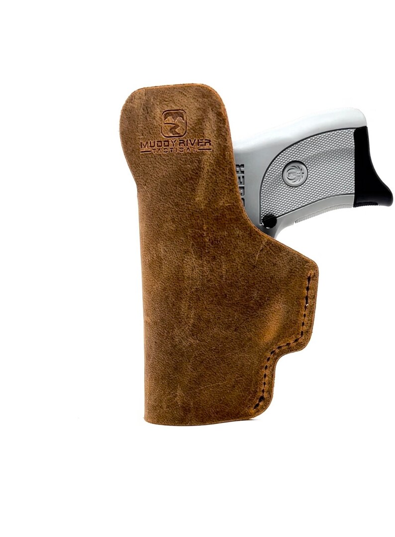 May include: A brown leather holster with a gun inside. The holster has the text "Muddy River Tactical" embossed on the side.