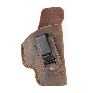 May include: A brown leather holster with a black metal clip. The holster has a distressed finish and visible stitching. The design is for concealed carry. The holster is made of leather.