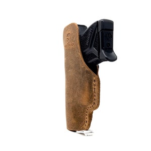 Puede incluir: Una funda de cuero marr&oacute;n con una pistola negra en su interior. La funda tiene la palabra "Hand River" grabada en relieve.