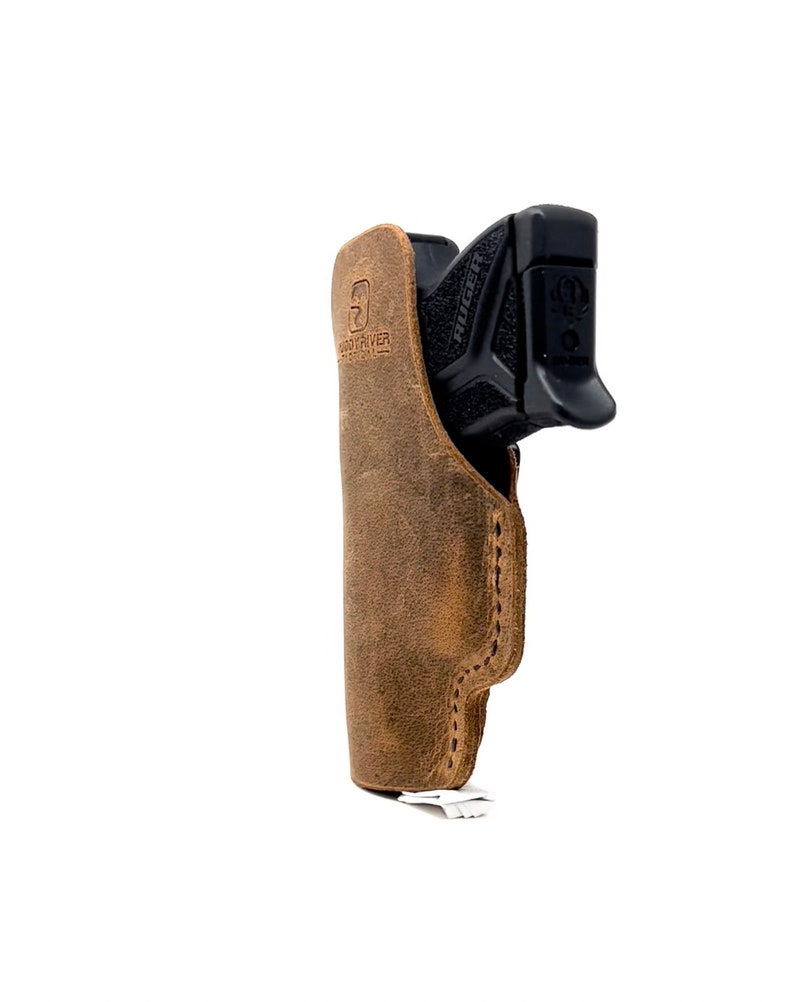 Puede incluir: Una funda de cuero marr&oacute;n con una pistola negra en su interior. La funda tiene la palabra "Hand River" grabada en relieve.