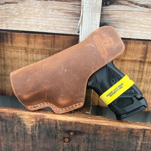 Puede incluir: Una funda de cuero marr&oacute;n para una pistola Ruger Lite Rack Security-9 negra. La funda est&aacute; hecha de cuero envejecido y tiene un borde cosido. La pistola tiene una pegatina amarilla en la empu&ntilde;adura que dice "Ruger Lite Rack Security-9".
