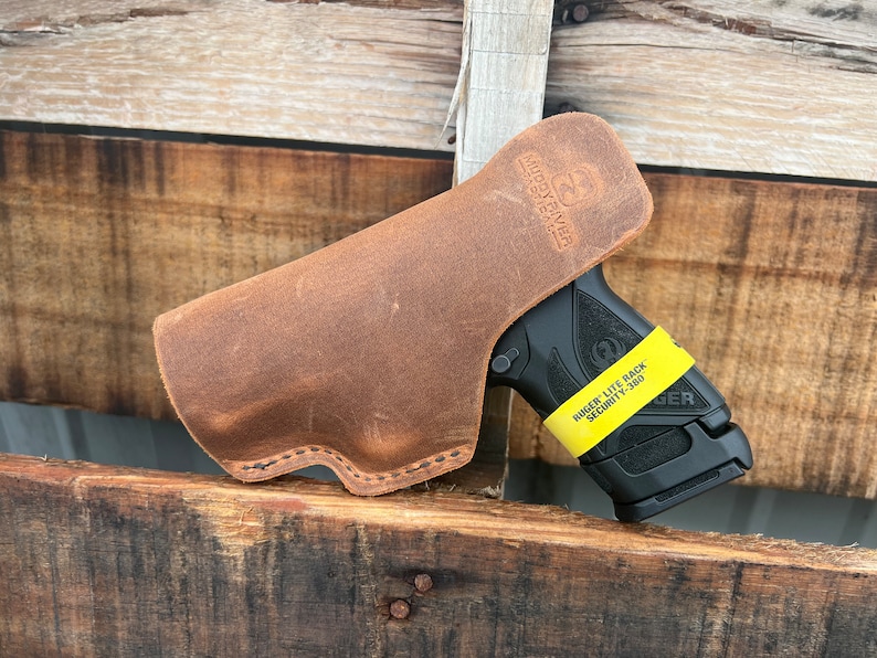 Puede incluir: Una funda de cuero marr&oacute;n para una pistola Ruger Lite Rack Security-9 negra. La funda est&aacute; hecha de cuero envejecido y tiene un borde cosido. La pistola tiene una pegatina amarilla en la empu&ntilde;adura que dice "Ruger Lite Rack Security-9".