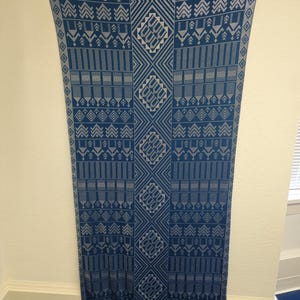May include: A long, rectangular textile with a repeating geometric pattern in shades of blue and white. The design features diamond, rectangular, and arrow-like shapes. The fabric is hanging vertically against a white wall.