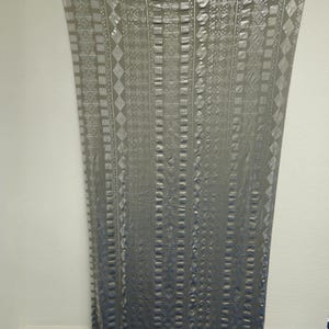 May include: A gray curtain with a geometric pattern of squares, rectangles, and diamonds. The curtain is hanging against a white wall. The fabric has a metallic sheen, and the design is embossed.